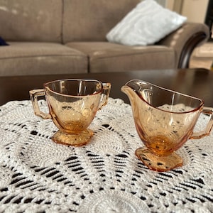 May include: A vintage peach-colored glass creamer and sugar bowl set. The creamer has a spout and handle, while the sugar bowl has two handles. Both pieces sit on a decorative, white crocheted doily.