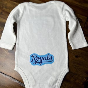 Pu&ograve; includere: Body da bambino bianco a maniche lunghe. Il body presenta la scritta "Royals" in blu sul fondo della schiena. Il testo &egrave; all'interno di un rettangolo arrotondato blu e bianco.