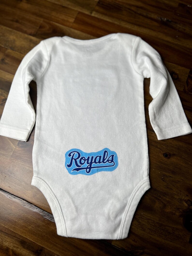 Pu&ograve; includere: Body da bambino bianco a maniche lunghe. Il body presenta la scritta "Royals" in blu sul fondo della schiena. Il testo &egrave; all'interno di un rettangolo arrotondato blu e bianco.