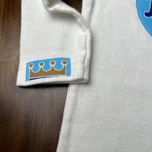 Pu&ograve; includere: Body da bambino bianco a maniche lunghe con toppa blu a forma di cuore. Il polsino della manica presenta un piccolo emblema a corona blu e oro. Il body &egrave; realizzato in materiale morbido.