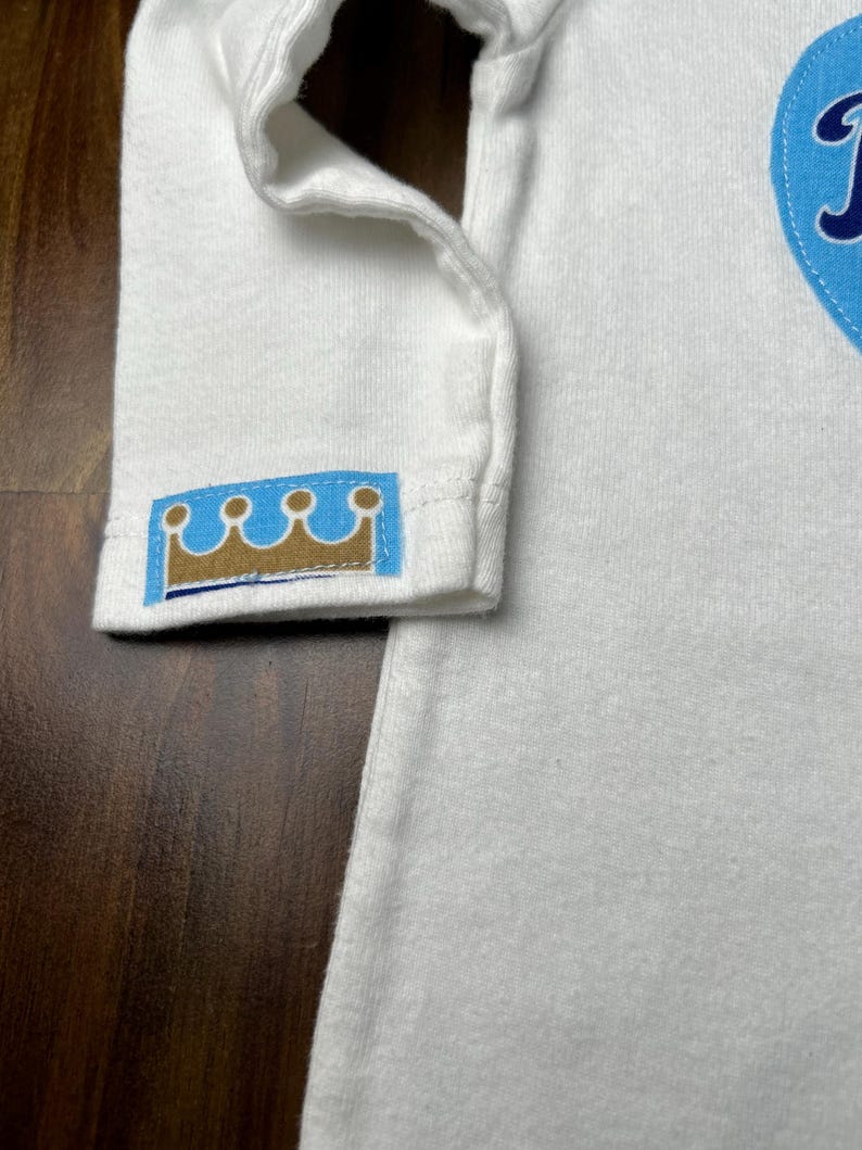 Pu&ograve; includere: Body da bambino bianco a maniche lunghe con toppa blu a forma di cuore. Il polsino della manica presenta un piccolo emblema a corona blu e oro. Il body &egrave; realizzato in materiale morbido.