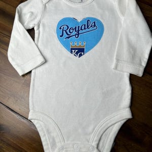 Pu&ograve; includere: Body da bambino bianco con maniche lunghe. Una grafica blu a forma di cuore al centro presenta la scritta "Royals", una corona e le lettere "KC". Il body &egrave; taglia 6.