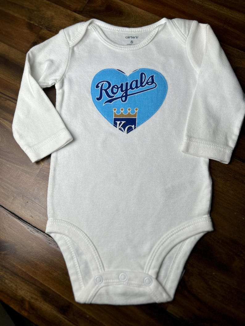 Pu&ograve; includere: Body da bambino bianco con maniche lunghe. Una grafica blu a forma di cuore al centro presenta la scritta "Royals", una corona e le lettere "KC". Il body &egrave; taglia 6.
