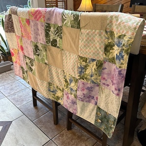 May include: A patchwork quilt draped over a dark wooden chair. The quilt features a variety of fabric squares in floral, polka dot, and solid patterns. Colors include pink, green, purple, and beige. The quilt appears handmade and adds a touch of warmth to the setting.