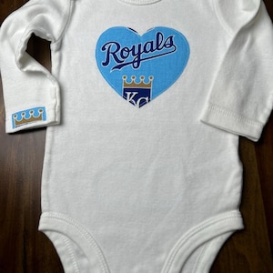 Pu&ograve; includere: Body da bambino bianco a maniche lunghe. Presenta una toppa blu a forma di cuore con la scritta "Royals" in corsivo e una grafica a corona. Il polsino della manica ha anche un piccolo disegno a corona. Taglia 6.