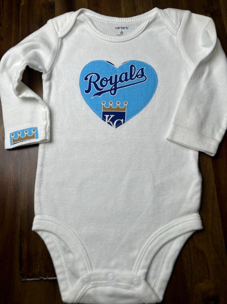 Pu&ograve; includere: Body da bambino bianco a maniche lunghe. Presenta una toppa blu a forma di cuore con la scritta "Royals" in corsivo e una grafica a corona. Il polsino della manica ha anche un piccolo disegno a corona. Taglia 6.