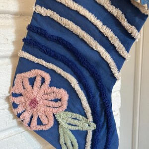 May include: A blue Christmas stocking with white, cream, and navy blue textured stripes. A pink and light green floral design is embroidered on the front. The top edge has a white, blue, and cream trim.
