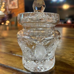 May include: A clear, cut-glass jar with a faceted, pointed lid. The jar has a bulbous body with intricate floral and geometric patterns. The lid and body are separate pieces. The jar is sitting on a wooden surface.