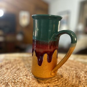May include: A tall ceramic mug featuring a tri-color glaze with a dark green top, a deep burgundy middle section with a dripping effect, and a textured golden-yellow base. The mug includes a curved handle that transitions from green to yellow.