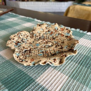 May include: A handmade ceramic dish with a unique, organic shape. The dish features a beige base with brown, blue, and orange speckled patterns. The rim has a wavy edge. The dish is placed on a green and white checkered tablecloth.
