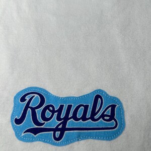 Pu&ograve; includere: Tessuto bianco con una toppa azzurra con la scritta "Royals" in blu scuro. La toppa ha un bordo azzurro e un bordo cucito.