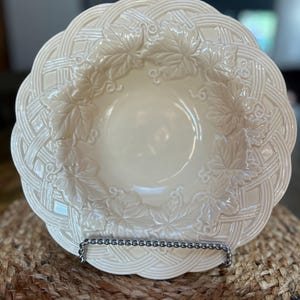 May include: Cream-colored ceramic serving bowl with a woven rim and embossed leaf design. The bowl is supported by a silver metal stand and sits on a woven mat. The bowl's interior is smooth and the rim has a detailed, textured pattern.