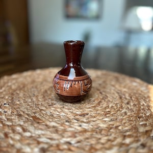 May include: A small, brown ceramic vase with a narrow neck and rounded body. The vase features a decorative band of carved geometric patterns in a lighter brown hue. The vase is displayed on a woven, circular mat.