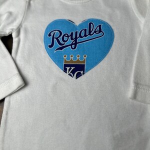 Pu&ograve; includere: Body da bambino bianco a maniche lunghe. Una toppa a forma di cuore azzurro chiaro &egrave; centrata sul petto, con la scritta "Royals" in blu navy. Sotto, una corona dorata e le lettere "KC" in blu navy.