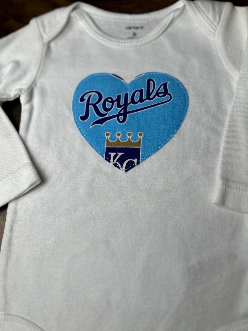 Pu&ograve; includere: Body da bambino bianco a maniche lunghe. Una toppa a forma di cuore azzurro chiaro &egrave; centrata sul petto, con la scritta "Royals" in blu navy. Sotto, una corona dorata e le lettere "KC" in blu navy.