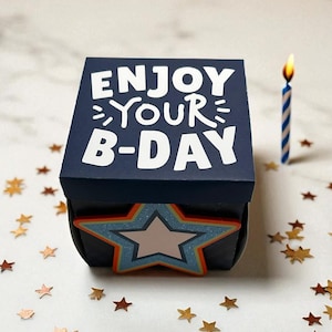 May include: A navy blue gift box with the text "ENJOY YOUR B-DAY" in white. A star-shaped embellishment is on the front. A lit blue and white striped candle stands next to the box. Gold star confetti is scattered around.