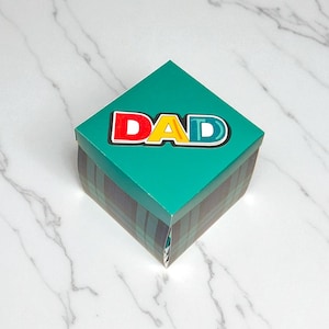 May include: Square gift box with a teal lid and plaid sides. The lid features the word "DAD" in red, yellow, and blue letters. The box is on a white marble surface.