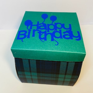 May include: A square gift box with a green lid and plaid sides. The lid features the words "Happy Birthday" in blue, along with blue balloon graphics. The box sides have a blue and green plaid pattern.
