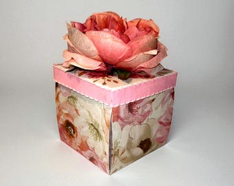 May include: A square gift box with a floral pattern in shades of pink, peach, and cream. A large, textured pink flower sits atop the box. The box has a scalloped pink border. The overall theme is feminine and decorative.
