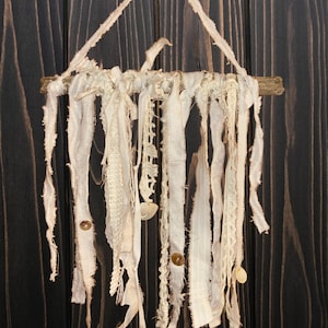 May include: A cream-colored wall hanging with a natural wood branch and hanging fabric strips. The strips vary in texture, including lace and frayed edges, and are adorned with small buttons and seashells. The background is a dark wood panel.