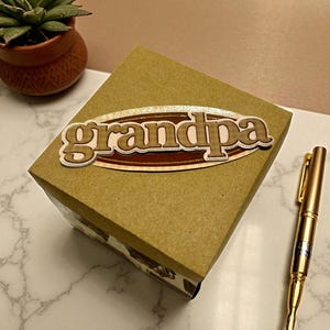 May include: A square gift box with a wooden "grandpa" embellishment. The box is a light brown color, and the text is in a brown, oval-shaped design. A gold pen rests on the marble surface next to the box. A small potted succulent is in the background.