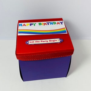 May include: A square gift box with a purple base and a red lid. The lid features a white rectangular label with the text "HAPPY BIRTHDAY" in multicolored letters above a rainbow graphic, and a smaller label reading "Let the Party Begin!"