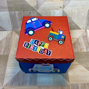 May include: A red box with a blue car, a blue truck, and a police car on the front. The box has the words "Happy Birthday" on the front in colorful letters.