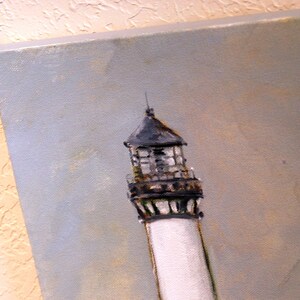 May include: A close-up of a white lighthouse with a black and gray roof and a black railing. The lighthouse is painted against a light blue sky.