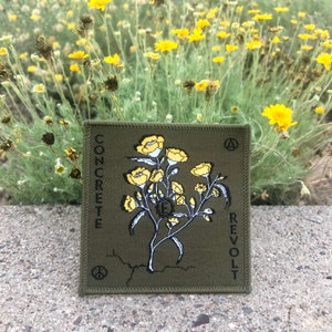 Pode incluir: Um remendo bordado verde com as palavras "CONCRETE REVOLT" e um símbolo de paz preto. O remendo apresenta um desenho estilizado de flores amarelas.