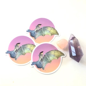 May include: Three round stickers with a pink and orange gradient background. Each sticker features a black and gray bat with yellow wings in flight.