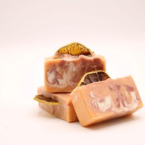 May include: Three bars of handmade soap with a swirl design in shades of pink and white. The top bar has a dried orange slice on top.