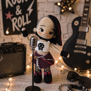 Pode incluir: Boneca de crochê de uma estrela do rock com cabelo preto e vermelho, vestindo um top branco com um gráfico de caveira, uma saia roxa e botas pretas. A boneca está segurando um microfone em frente a uma guitarra preta e um amplificador. Uma placa que diz "Rock & Roll" está no fundo.