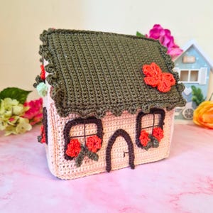 Könnte beinhalten: Ein gehäkeltes Haus in zartem Rosa mit einem dunkelgrünen Dach und dekorativen roten Blumen. Die Fenster und die Tür sind braun umrandet. Das Dach hat einen gewellten Rand, und ein roter Schmetterling ist am Dach befestigt.