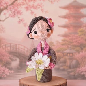 May include: A crochet doll with dark brown hair styled in a bun, wearing a dark grey garment with a pink collar and a yellow sash. The doll has rosy cheeks and holds a large white flower with a yellow centre and green leaves in its hands.