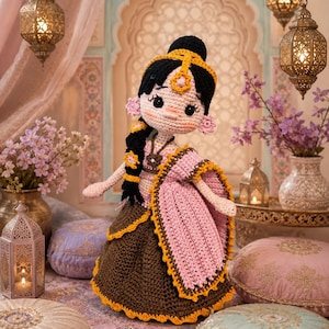 May include: A handcrafted crochet doll featuring black hair styled in a braid with yellow floral accents, wearing a pink and brown outfit with yellow trim. The doll has large black eyes, pink cheeks, and a yellow headpiece with a central gem detail.