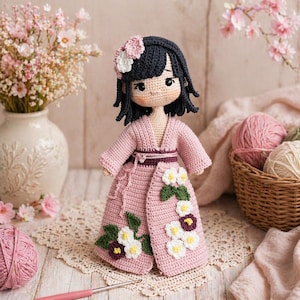 Peut inclure: Poupée au crochet faite à la main avec des cheveux noirs et une robe rose de style kimono. La poupée a un accessoire de cheveux en forme de fleur et est ornée d'appliqués de fleurs blanches et violettes. La poupée mesure environ 25 cm de haut.