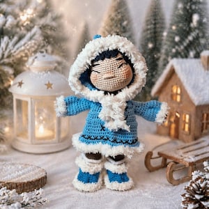Op de afbeelding: Een handgemaakte gehaakte pop gekleed in een blauwe winteroutfit met witte rand en een donzige capuchon. De pop staat in een besneeuwde scène met een lantaarn, een miniatuurhuis en een slee op de achtergrond. Het gezicht van de pop is bruin.