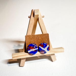 May include: A pair of round, blue, red, and white patterned stud earrings displayed on a wooden easel.