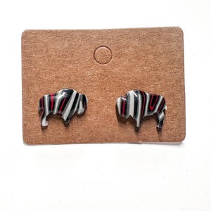 May include: A pair of stud earrings featuring a bison design. The earrings are black, white, and red, with a striped pattern. They are displayed on a brown card.