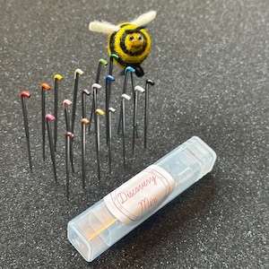 May include: A collection of sewing pins with colorful heads, arranged with a felted bee figurine. The pins are in a clear plastic case labeled "Discovery Mix". The bee is yellow and black with white wings.