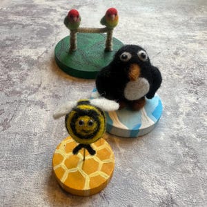 Peut inclure: Trois figurines en laine feutrée sur des socles en bois circulaires : deux petits oiseaux rouges et verts sur un socle vert, un manchot noir et blanc sur un socle bleu et blanc, et une abeille rayée jaune et noire sur un socle à motif alvéolé.