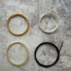 Puede incluir: Cuatro mechones de cabello circulares en tonos rubios, blancos y negros. El cabello está dispuesto sobre una superficie texturizada de tonos neutros, mostrando los diferentes colores y texturas del cabello.