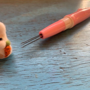 May include: A pink felting tool with multiple needles and a white felt bunny holding a carrot. The bunny is sitting on a blue wooden surface.