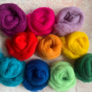 Puede incluir: Diez ovillos de lana en varios colores, incluyendo morado, rosa, rojo, naranja, azul verdoso, azul, verde y verde oscuro. Los ovillos están dispuestos en semicírculo sobre una superficie gris claro.