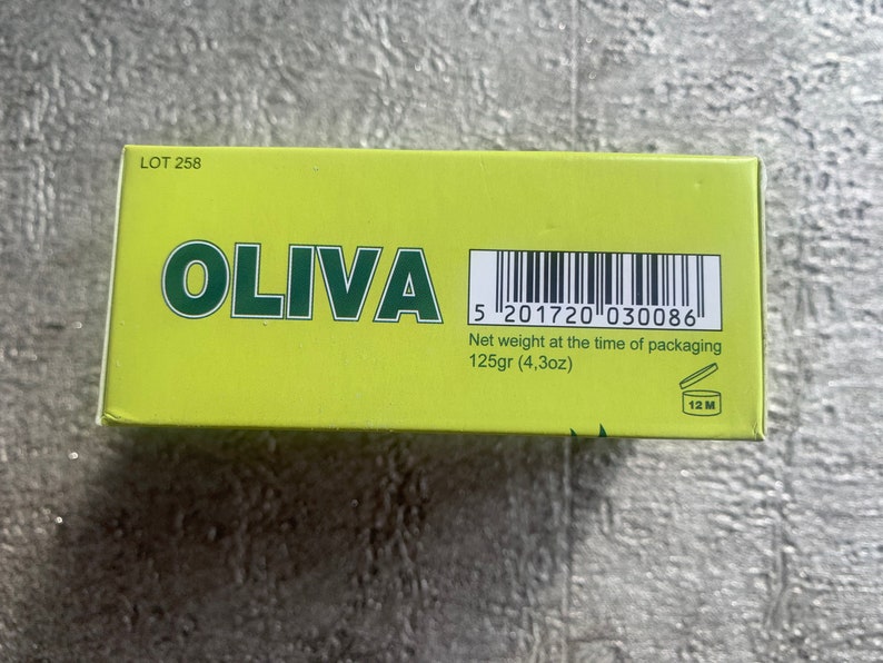 May include: A yellow box with the word "OLIVA" in green letters. The box has a barcode and the text "Net weight at the time of packaging 125gr (4,3oz)". There is also a symbol of an open jar with the text "12 M" inside.