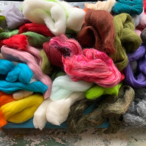 Op de afbeelding: Een assortiment van kleurrijke wolroving in verschillende tinten groen, blauw, roze, rood, oranje, geel, bruin, wit, grijs en paars. De wol is op een blauwe ondergrond in een stapel gelegd.