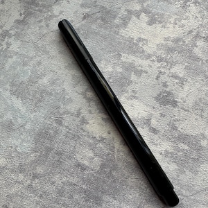 Può includere: Un pennarello nero con punta arrotondata. La penna è posizionata in diagonale su una superficie grigia strutturata. La finitura lucida della penna contrasta con lo sfondo opaco.