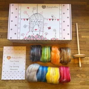 Puede incluir: Un kit de manualidades que incluye una caja blanca con una ilustración de jaula y el texto heidi feathers, ocho manojos de lana cardada en gris, verde, naranja, marrón, blanco, azul, amarillo y rosa, un huso de madera y una hoja de instrucciones.