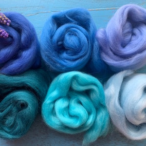 May include: Six balls of wool in shades of blue and teal. The wool is arranged in a semi-circle on a blue wooden surface.