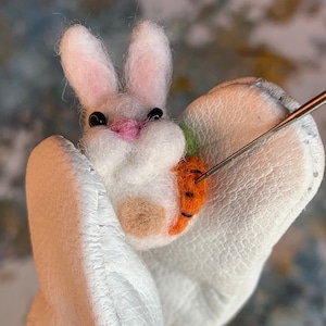 May include: A white felt bunny with pink ears and black eyes holding a small orange carrot. The bunny is being sewn onto a white felt background with a needle and thread.